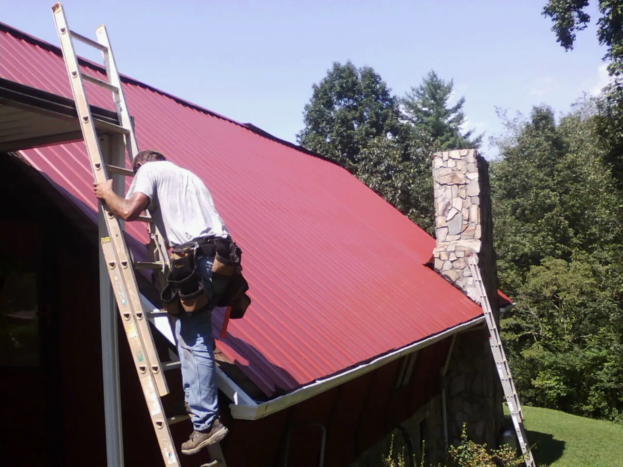 Roofer on a ladder beside a red metal roof and stone chimney — Soffit & Fascia Repair by Roofing Brothers in Chapmansboro